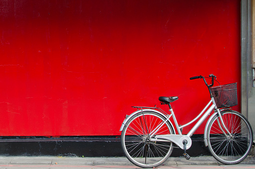 Silver bike with basket in the front parking in front of a red wall. 