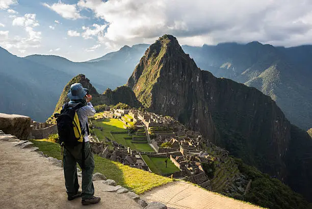 Tourist photographing Machu Picchu illuminated by the last sunlight from above with smartphone. Scenic sky with clouds and sun rays. Concept of sharing travel moments using technology.