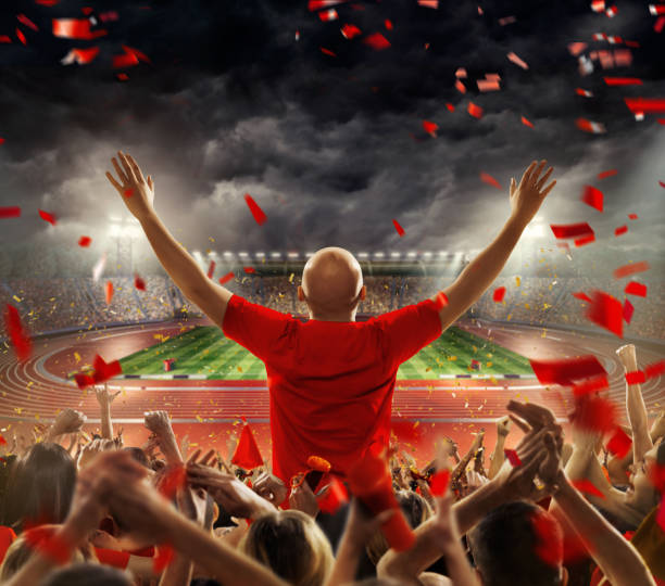Fans at . stadium with running tracks On the foreground a group of cheering fans watch a sport championship on stadium. One man stands with his hands up to the sky. People are dressed in red colors. A long-range shot of a stadium field, floodlights and seating. A green field, with painted white lines, is visible in the foreground. In the background are diffuse out-of-focus stadium seats. Large, bright floodlights are in the top-left and top-right corners of the stadium. track and field fans stock pictures, royalty-free photos & images