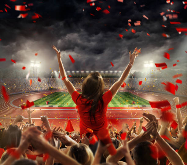 Fans at . stadium with running tracks On the foreground a group of cheering fans watch a sport championship on stadium. One girl stands with his hands up to the sky. People are dressed in red colors. A long-range shot of a stadium field, floodlights and seating. A green field, with painted white lines, is visible in the foreground. In the background are diffuse out-of-focus stadium seats. Large, bright floodlights are in the top-left and top-right corners of the stadium. track and field fans stock pictures, royalty-free photos & images