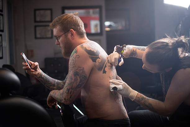 Artist tattooing man's back in studio A photo of artist tattooing man's back. Tattoo expert is working in studio. Shirtless male customer is using smart phone while getting his back tattooed. body adornment rear view young men men stock pictures, royalty-free photos & images