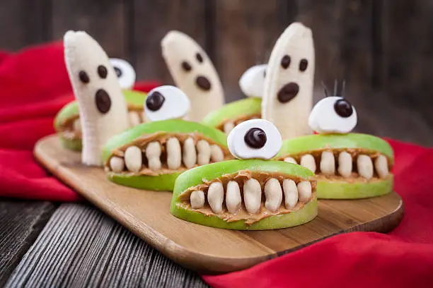 Funny halloween eadible monsters scary food healthy vegetarian snack dessert recipe for party decoration. Homemade spooky cyclop apples with teeth and banana ghosts on vintage wooden background. Natural treat
