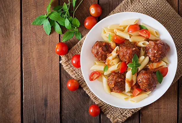 Penne pasta with meatballs in tomato sauce in a white bowl. Top view