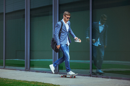 Young businessman commuting to work skateboarding. The man is casually dressed and wears sunglasses, earphones and carries black briefcase hung on shoulder.