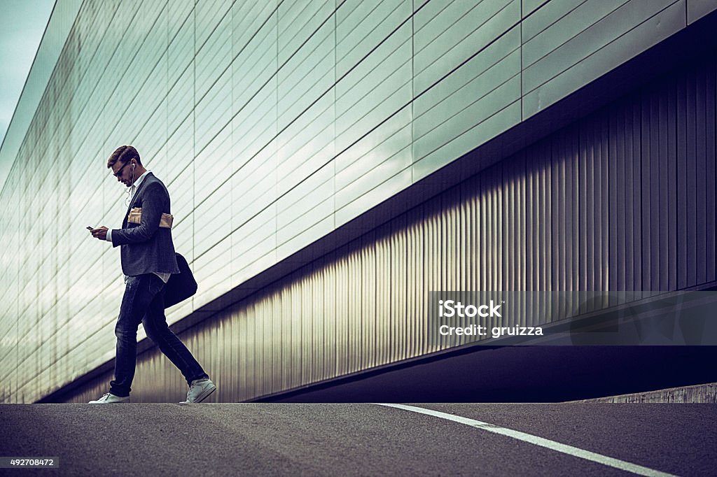 Young casual businessman using smartphone in the urban environment Young businessman using smartphone in the urban environment The man is casually dressed and wears sunglasses, earphones and carries black briefcase hung on shoulder. Walking Stock Photo