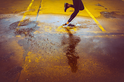 Young person running over the yellow parking lot. Black sport clothing - sport shoes, running tights, and a jacket. High angle view of a runner's legs and its reflection in the water.