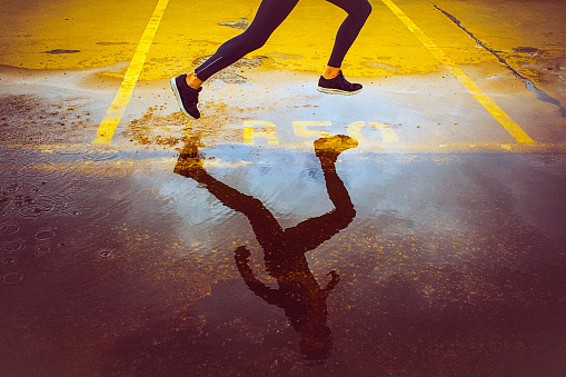 Young person running over the yellow parking lot. Black sport clothing - sport shoes, running tights, and a jacket. High angle view of a runner's legs and its reflection in the water.