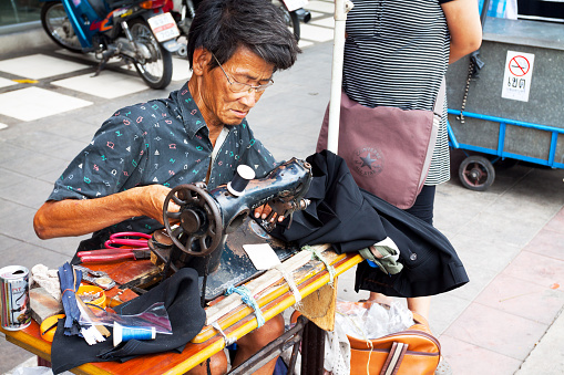 Bangkok,Thailand - May 31, 2013: A mature male thai tailor is working and sewing on sidewalk in Bangkok. Scene is in Bang Khen on Pahonyothin Road. A woman is standing next to men.