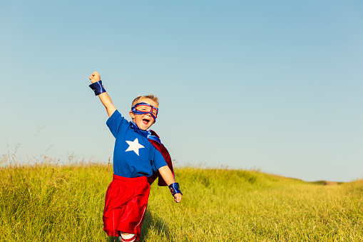 A young boy wearing a superhero cape and mask is ready to face life's challenges with confidence. He has his arm raised to the sky while standing in tall grass. Image taken in Wiltshire, United Kingdom.