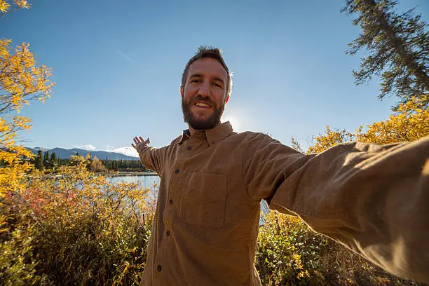 Young cheerful man traveling in Canada takes a selfie portrait by the lake. Autumn season.