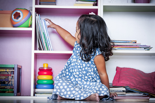 Child read, cute little girl selecting a book on bookshelf and sitting on floor