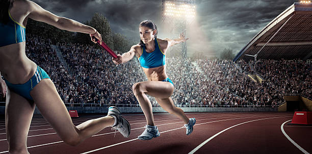Running Pass on . Stadium A woman athletes sprinting and passing race baton on the track on the dramatic evening . stadium. The bleachers are full of spectators. The sky is dark and cloudy. The woman is wearing an unbranded bra top with small bottoms. relay photos stock pictures, royalty-free photos & images