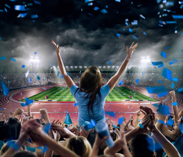 Fans at . stadium with running tracks On the foreground a group of cheering fans watch a sport championship on stadium. One girl stands with his hands up to the sky. People are dressed in blue colors. A long-range shot of a stadium field, floodlights and seating. A green field, with painted white lines, is visible in the foreground. In the background are diffuse out-of-focus stadium seats. Large, bright floodlights are in the top-left and top-right corners of the stadium. track and field fans stock pictures, royalty-free photos & images