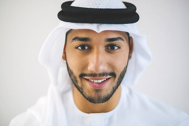 Horizontal Portrait of Young Smiling Arab Man Young man from United Arab Emirates is posing for a close-up portrait looking at the camera and smiling charmingly. Wearing traditional white clothing: kandura (also known as dish dash), agal and kaffiyeh. Dark brown eyes, black facial hair. Background of the image is light matte grey. Image was made in Dubai, United Arab Emirates. arabian peninsula photos stock pictures, royalty-free photos & images