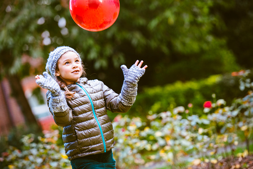 Young girl playing with red ball in a park. She is wearing warm clothes. Shot on a cold autumn day.