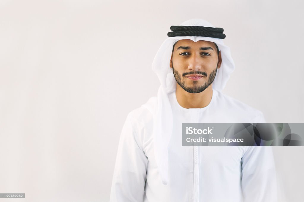 Content Young Arab Man in Traditional Clothing Young and confident Emirati man is standing in front of light grey wall, hands let down, looking at the camera smiling lightly. Model's clothing is white and black making good contrast with his face. Image contains copy space on the left. Made in Dubai, United Arab Emirates. Men Stock Photo