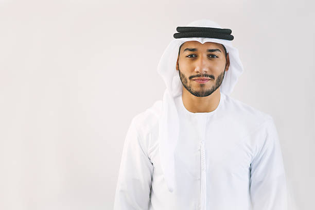 Content Young Arab Man in Traditional Clothing Young and confident Emirati man is standing in front of light grey wall, hands let down, looking at the camera smiling lightly. Model's clothing is white and black making good contrast with his face. Image contains copy space on the left. Made in Dubai, United Arab Emirates. arabian peninsula photos stock pictures, royalty-free photos & images