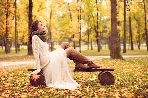 Beautiful girl sitting on a bench in park and enjoy while watching autumn colors all around. He's wearing a white skirt, black leather boots, handmade sweater and green scarf. She is relaxed, happy and looks into the distance.