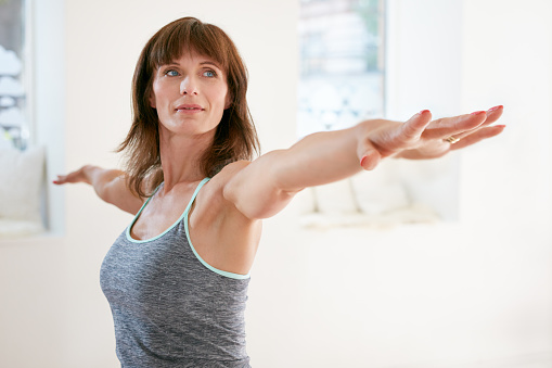 Portrait of mature woman stretching her arms and looking away at gym. Beautiful woman performing yoga in warrior pose, Virabhadrasana.