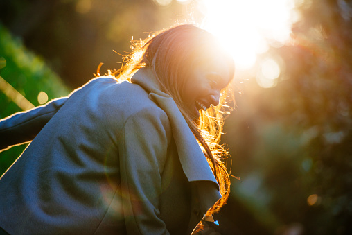 Young woman, enjoying outdoors at sunset on a sunny autumn day. She is wearing warm clothing. She is smiling and having fun. Sun is shining from back.