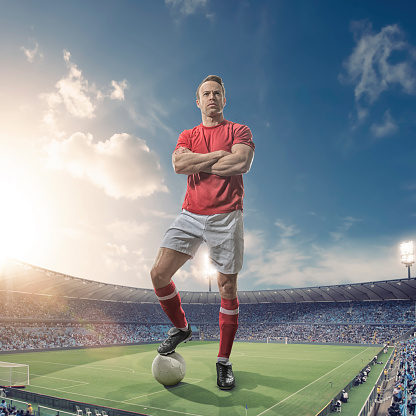 A conceptual image of a giant male footballer standing with arms folded and foot on football, looking into the distance, in a generic soccer stadium full of spectators. The huge man is wearing red and white soccer kit, and towers above the floodlit stadium under a dusky evening sky.