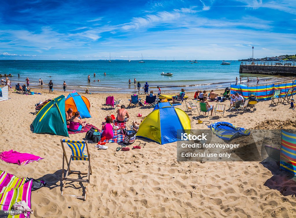 Crowds sunbathing on sandy ocean beach with colourful tents England Swanage, UK - August 1, 2015: Families enjoying the summer sunshine on deckchairs on the sandy beach at the popular seaside holiday resort of Swanage in Dorset, UK. Composite panoramic image created from four contemporaneous sequential photographs. UK Stock Photo