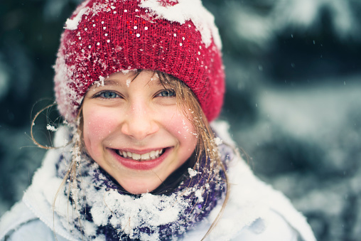 Winter portrait of a beautiful little girl laughing and having a lot of fun in snow. The girl aged 8 and her scarf, face and cap are snow covered. Some of the snow have melted on her cheeks.