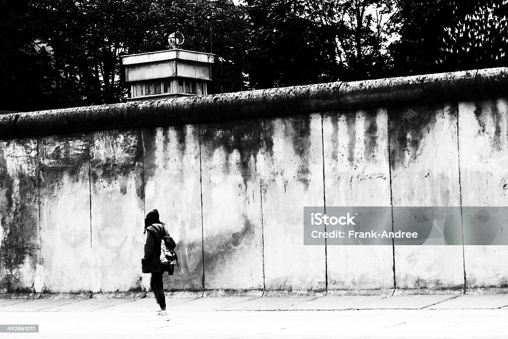 Young woman goes to the Berlin Wall along Black / white photograph of the Berlin Wall. A young woman walks carelessly along the wall. Behind the Berlin Wall can be seen, a former watchtower. Reminder of the division of Berlin. Former border fortification in Berlin. Fall Of The Berlin Wall Stock Photo