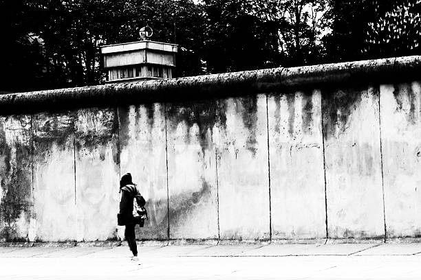 Young woman goes to the Berlin Wall along Black / white photograph of the Berlin Wall. A young woman walks carelessly along the wall. Behind the Berlin Wall can be seen, a former watchtower. Reminder of the division of Berlin. Former border fortification in Berlin. cold war photos stock pictures, royalty-free photos & images