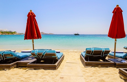 Sun beds with couchins, umbrellas, golden sand and the blue sea in Mykonos, Greece. A greek island summer holiday scene at the Psarou beach with a boat in the crystal clear water.