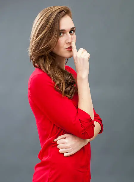secret and taboo concept - mysterious 20s woman wearing red shirt asking for silence with finger on mouth,studio shot