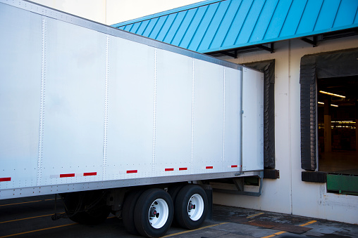 White long semi trailer with an open door stand close in the dock warehouse with a green sloping roof to unload or load cargo for delivery and transport to safety labels on the walls of the semi trailer.