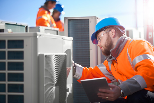 An air conditioning engineer is finishing the installation of several units on a rooftop. Two colleagues can be seen also installing units in the background. They are wearing hi vis jackets, hard hats and safety goggles.