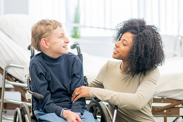 Caregiver Talking to Little Boy A little boy is sitting in his wheelchair with his caregiver. They are sitting together in the hospital and are smiling. paraplegic stock pictures, royalty-free photos & images