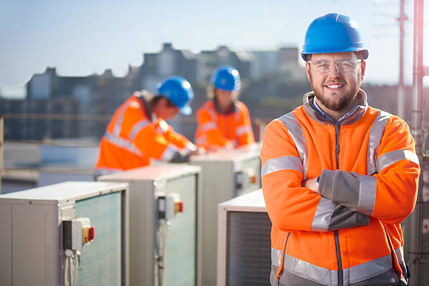 air conditioning engineer portrait An air conditioning engineer is finishing the installation of several units on a rooftop. Two colleagues can be seen also installing units in the background. They are wearing hi vis jackets, hard hats and safety goggles. maintenance engineer photos stock pictures, royalty-free photos & images