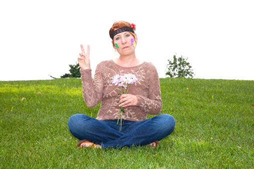 Mature woman dressed as a sixties flower child sitting in the grass holding daisies and flashing a peace sign