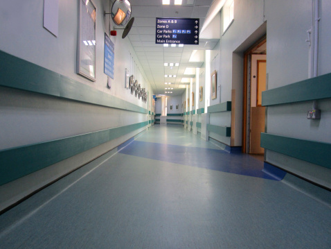 Photo showing a colourful hospital corridor, with turquoise and blue vinyl flooring, a series of small rectangular windows and a row of strip lights within the ceiling tiles. Various medical treatment rooms, nurses' stations, offices of doctors and hospital wards lie off this corridor, which is usually extremely busy. This picture was taken very early in the morning.