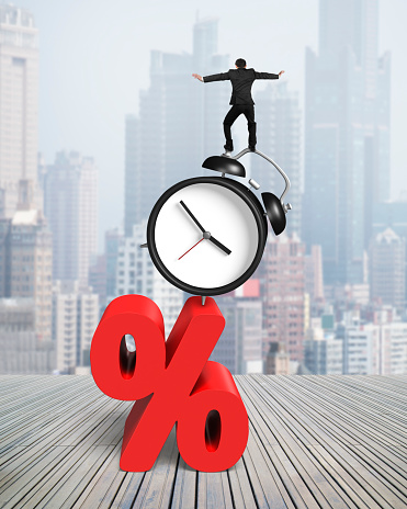 Businessman balancing on alarm clock and red percentage sign, on cityscape and wood floor background.