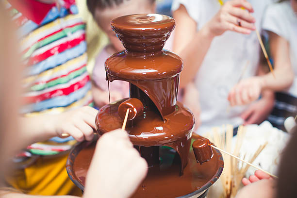 Chocolate Fountain With Fondue, Fruits and Marshmallow on children party Vibrant Picture of Chocolate Fountain Fontain on childen kids birthday party with a kids playing around and marshmallows and fruits dip dipping into fountain drinking fountain stock pictures, royalty-free photos & images