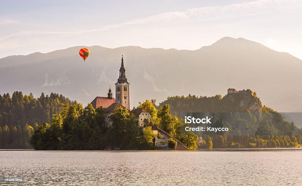 Church in Lake and Castle, Slovenia with Hot Air Balloon Detail of Famous Catholic Church on Little Island in Bled Lake and Bled Castle on a Rock in Slovenia with Hot Air Balloon Flying with Mountains in Background at Sunrise 2015 Stock Photo