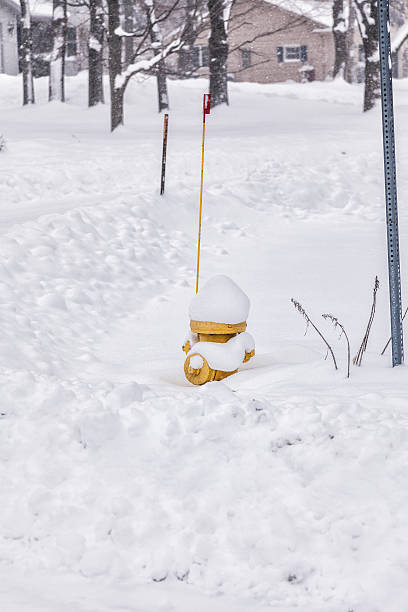 A yellow fire hydrant being buried by a winter blizzard snow storm at a suburban neighborhood street corner. The red tipped flag is attached to ensure emergency fire fighter responders can find the hydrant quickly even if covered by deep snow. The thin metal rod to the left is placed there to prevent snowplow truck drivers from cutting the street corner too tightly when the edge of the asphalt is buried.
