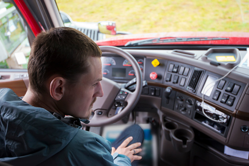 The truck driver sitting in the cab of modern comfort and ergonomic semi truck behind the wheel and interior dashboard with numerous control buttons and switches. The driver of semi truck assessment the situation, looking in the right rearview mirror.