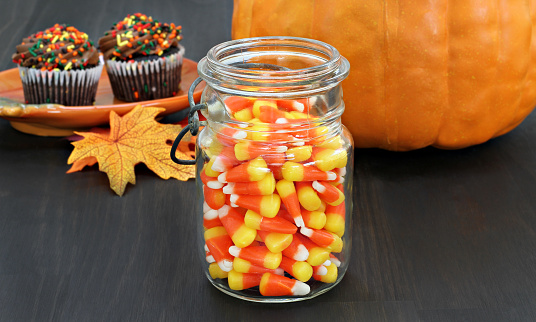 A jar of candy corn in a rustic mason jar.  Autumn background with a pumpkin, leaves and fall cupcakes.