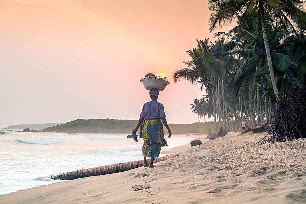 African woman walking on beautiful beach Anomabo, Ghana - March 3, 2014: African woman with heavy load on her head going home, toward the Sun west africa coast photos stock pictures, royalty-free photos & images
