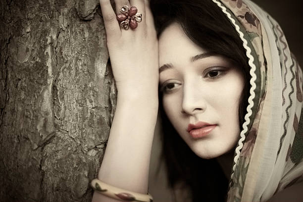 Hand tinted retro style outdoor image of Beautiful Sad Indian young woman standing by holding and resting with tree trunk. She is wearing dupatta. Horizontal composition with copy space and selective focus.