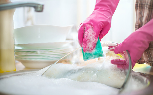 Closeup of unrecognizable person washing big glass serving bowl in kitchen sink. She's wearing pink gloves and using green sponge and dishwashing detergent. The sink is full with soap sud. Blurry plates on background. Side view,low angle shot.