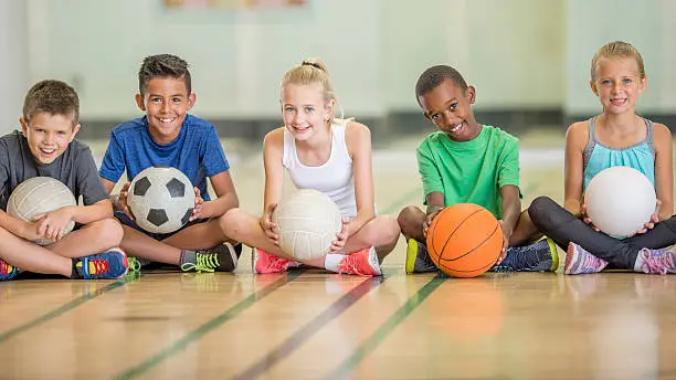 A multi-ethnic group of elementary age children are sitting in a row on the basketball court - they are smiling and looking at the camera.