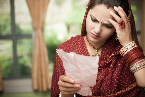 Indoor day time image of a beautiful, newly married Indian young, worried and disturbed woman from a wealthy family standing against a window background by holding two torn wrinkled pieces of paper to which she is recombining and reading and also holding her head out of worry and headache. She is in traditional Hindu dress that is Salwar Kameez, dupatta and bangles. Horizontal composition with copy space and selective focus.
