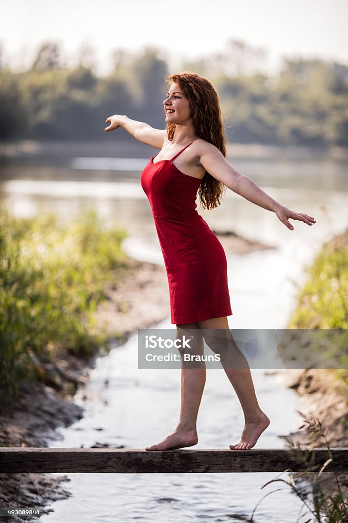 Happy woman balancing on wooden post above the stream. Young redhead woman walking on wooden post above the stream while balancing with her arms outstretched. 2015 Stock Photo