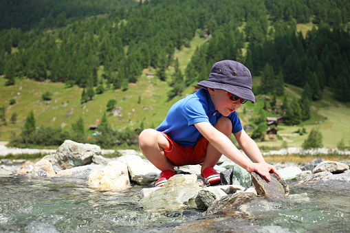 A little boy is playing in a mountain river on a summer day as he is building a water dam with stones and rocks. This image is taken in Zinal valley in Valais region Switzerland.